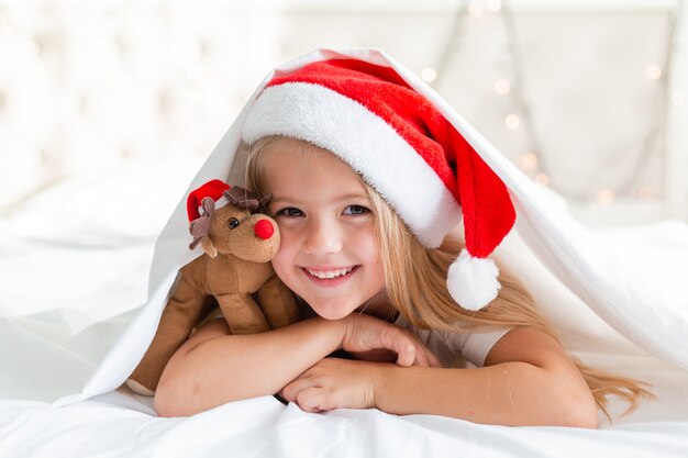 closeup retrato de uma menina loira deitada na cama com um chapéu de Papai Noel e um cervo de brinquedo