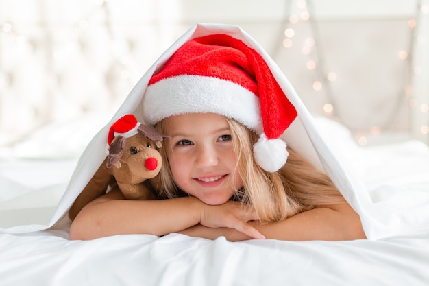 closeup retrato de uma menina loira deitada na cama com um chapéu de Papai Noel e um cervo de brinquedo