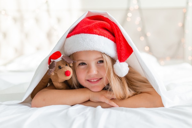 Closeup retrato de uma menina loira deitada na cama com um chapéu de papai noel e um cervo de brinquedo