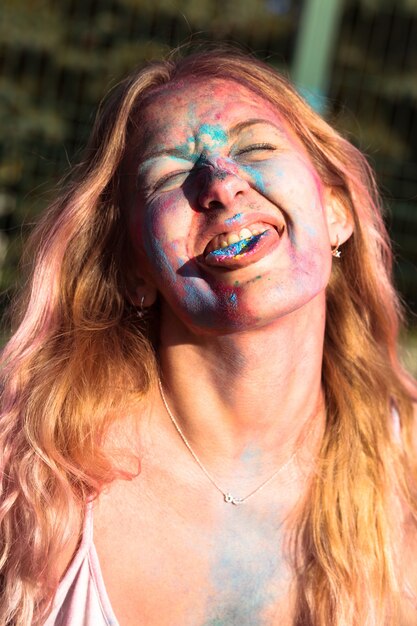 Closeup retrato de uma mulher loira alegre coberta com tinta colorida Holi, mostrando a língua