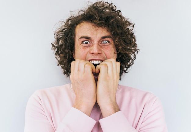 Closeup retrato do jovem modelo masculino com cabelo encaracolado com expressão assustada e ansiosa morde unhas usa suéter rosa com medo expressões faciais humanas e conceito de emoções negativas