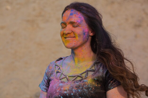 Closeup retrato do modelo moreno positivo posando coberto com tinta Holi seca no deserto. Espaço para texto