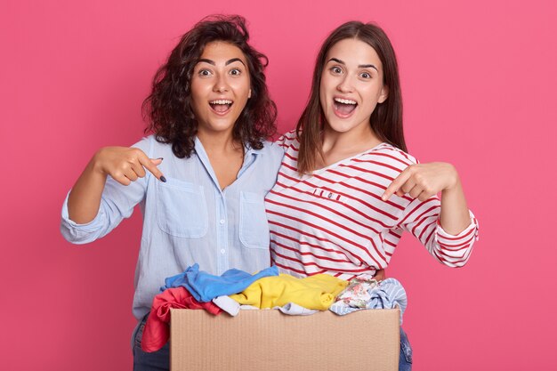 Closeup retrato de dos chicas emocionadas apuntando a la caja de cartón con ropa para usar secundaria