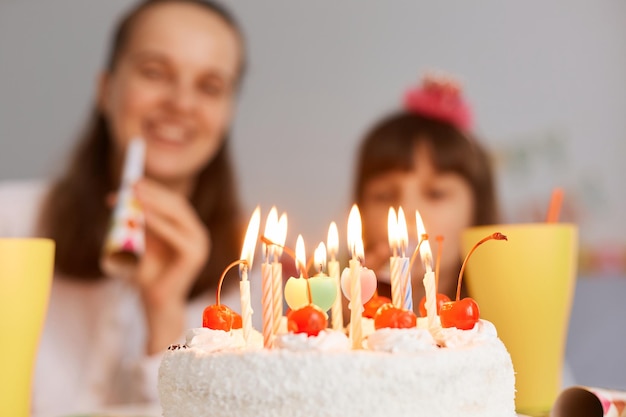 Closeup retrato de gran pastel de crema blanca con velas, delicioso postre, madre con cuerno de fiesta y niña en el fondo, gente celebrando cumpleaños en casa.