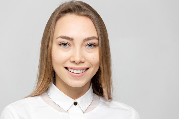 Closeup retrato de hermosa mujer rubia atractiva joven feliz en una camisa blanca con la piel limpia mirando a la cámara contra el fondo grisblanco