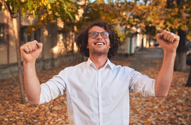 Foto closeup retrato horizontal de estudante do sexo masculino vencedor feliz ou empresário com cabelo encaracolado sorrindo com punhos de expressão vencedora bombeado posando na rua outono copie o espaço sucesso e triunfo