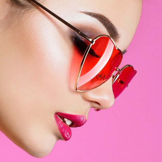 Closeup retrato de joven hermosa mujer roja con gafas de sol en forma de corazón. Ojos ahumados y labios rojos