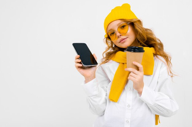 Closeup retrato de una linda chica adolescente caucásico en un gorro de invierno y bufanda con un teléfono con una placa en la mano y un vaso de papel en blanco