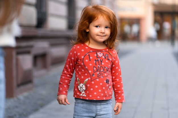 Closeup retrato de lindo adorable sonriente niña caucásica pelirroja de pie en el parque de otoño otoño afuera, mirando a puerta cerrada, concepto de infancia feliz estilo de vida