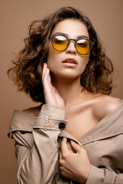 Closeup retrato de modelo de moda de belleza con piel limpia y cabello rizado en estiramiento de capa de biege, modelo con gafas de moda, hombros desnudos