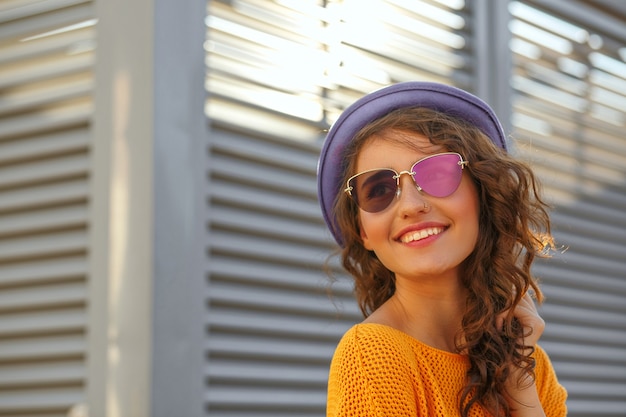 Closeup retrato de modelo morena muy sonriente con pelo rizado con gafas y sombrero azul. Espacio para texto