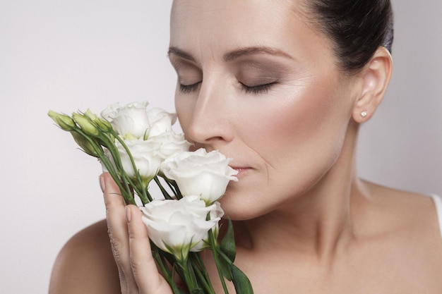 Closeup retrato de mujer hermosa alegre con piel perfecta y arrugas envejecidas con olor a flores. Concepto de cuidado de la piel, cosméticos de envejecimiento, aislado sobre fondo gris, espacio de copia, foto de estudio, interior