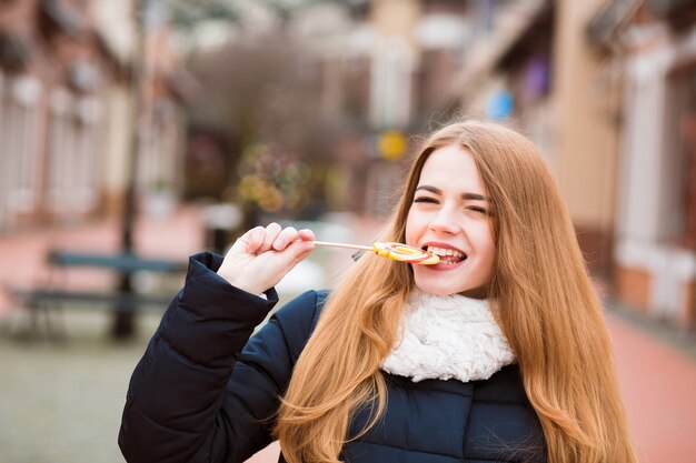 Closeup retrato de mujer rubia alegre comiendo coloridos dulces navideños en el fondo de la calle