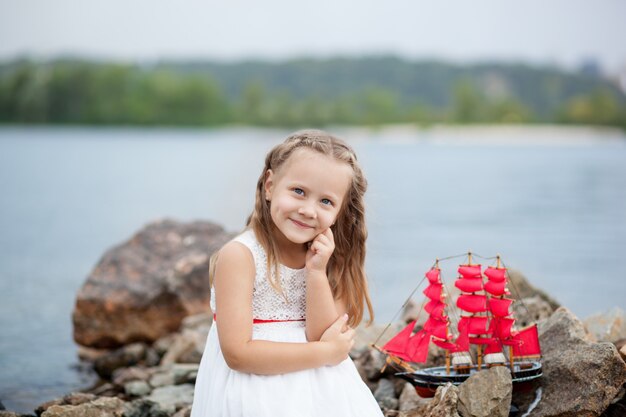 Closeup retrato niña linda en vestido blanco y velas rojas. Un niño sentado en el mar con barco de juguete. Concepto de infancia. Juegos de arena en la playa. Concepto de descanso El niño juega con el juguete al aire libre