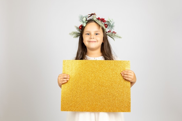 Closeup retrato de niña sonriente encantadora en corona de navidad sosteniendo banner en blanco brillante dorado isol ...