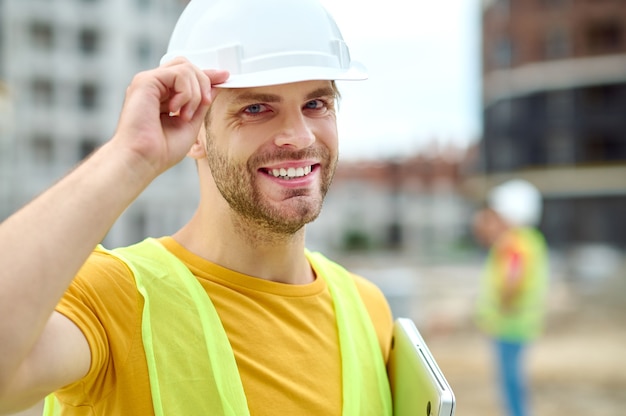 Closeup retrato de un sonriente ingeniero masculino lindo alegre con un portátil en una mano tocando su casco