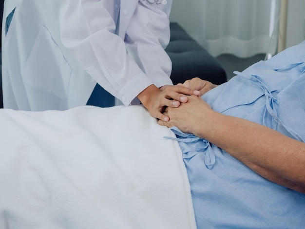 Closeup Ärztin im weißen Anzug hält sich an der Hand der älteren Seniorin in hellblauem Kleid mit einem Kochsalzschlauch, der auf dem Krankenhausbett liegt und das Patientenkonzept fördert