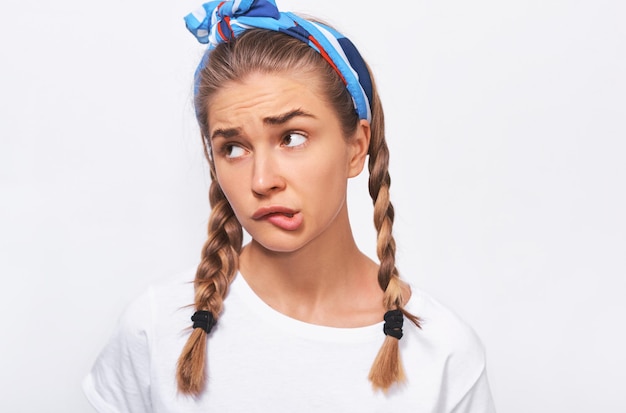 Foto closeup studio portrait von nachdenklich blonde schöne junge frau trägt wghite t-shirt blaues stirnband und zöpfe frisur sieht unsicher posen vor weißem hintergrund menschen emotionen konzept