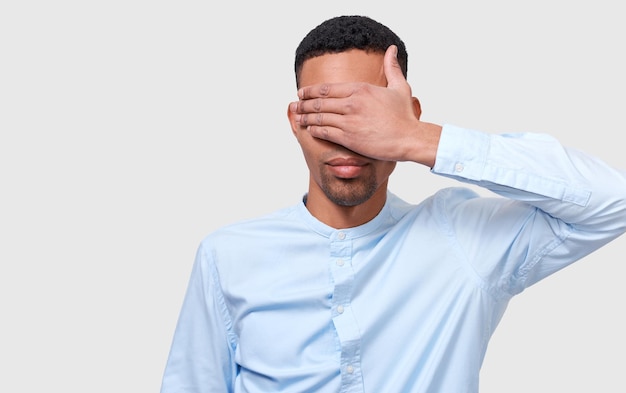 Closeup Studioportrait eines gut aussehenden afroamerikanischen Mannes bedecken Augen mit der Hand, die das blaue beiläufige Hemd trägt, das auf weißem Hintergrund lokalisiert wird Geheimhaltung Geschäftsmann mit verbundenen Augen Schutz Menschen und Unternehmen