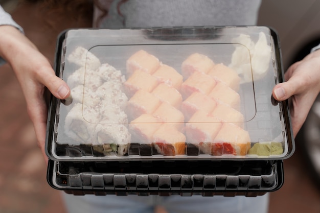 Closeup sushi en caja servicio en línea de entrega de alimentos saludables. La niña tiene 2 juegos de sushi en las manos. Rollos de cocina japonesa, salsa de soja, wasabi.