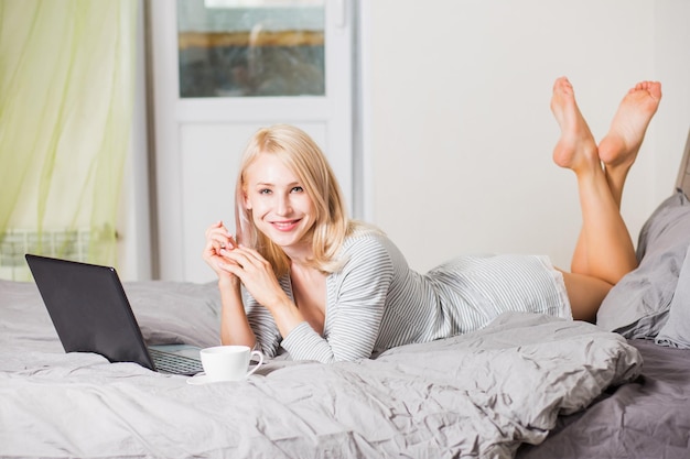 Closeup tiro da blogueira loira bonita caucasiana com sorriso positivo, deitada na cama, usando o computador laptop e a conexão de alta velocidade à Internet, relaxando no quarto no fim de semana.