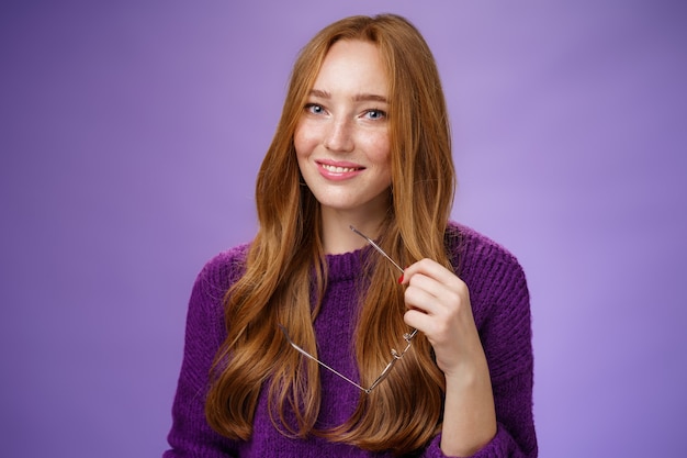 Closeup tiro de encantadora mulher ruiva macia e bonita de suéter roxo segurando óculos e sorrindo.