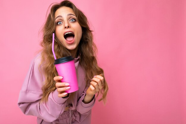 Closeup tiro de foto de uma bela jovem loira espantada, vestindo roupas esporte rosa isoladas sobre um fundo rosa, segurando uma xícara de café de papel para maquete bebendo e olhando para a câmera com a boca aberta