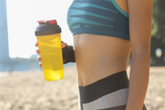 Closeup tiro de mulher de aptidão com formas de esportes, segurando a garrafa de água, posando na praia. Espaço para texto