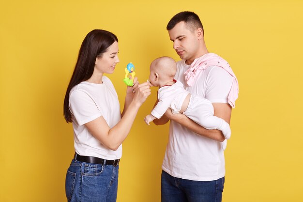 Closeup tiro do pai acariciando o bebê nas mãos do pai e a mãe ao lado deles dando chupeta para o bebê e mostrando o brinquedo para o recém-nascido, família vestindo roupas casuais e pose contra a parede amarela.