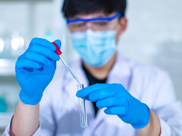 Closeup tiro facial do jovem cientista masculino profissional asiático em jaleco branco usando óculos de proteção de segurança usando lentes de microscópio olhando zoom na amostra de microbiologia na placa de vidro.