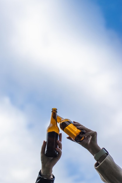 Closeup vertical de amigos brindando com garrafas de cerveja nas mãos contra o céu