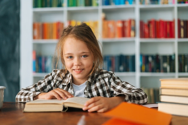 Closeup vista frontal do aluno alegre aluno da escola fazendo lição de casa lendo livro de papel sentado no