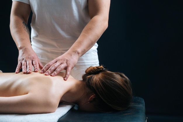 Closeup vista lateral do massagista masculino massageando as costas de uma jovem deitada na mesa de massagem em fundo preto Fêmea com pele perfeita recebendo massagem relaxante Conceito de tratamentos de spa de massagem