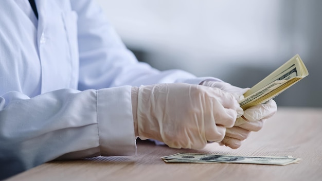 Closeup weibliche Hände in weißen Latexhandschuhen Apotheker Arzt Wissenschaftler im medizinischen Kittel sitzt am Tisch mit einer Flasche Pillen Medikament zählen Geld Banknoten Dollar Bargehalt Gewinne aus dem Verkauf von Medikamenten