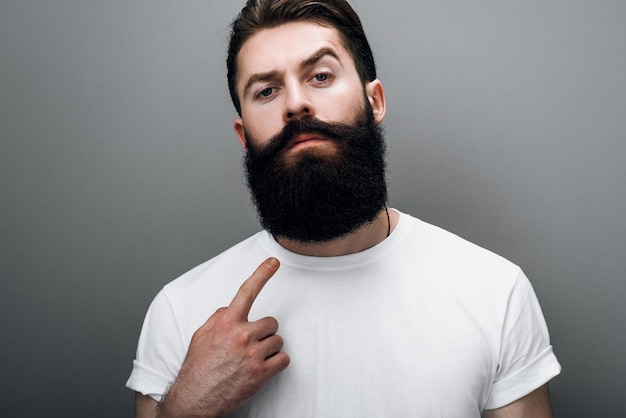 Clsoe up retrato recortado de brutal hombre caucásico guapo indica su barba de moda posando sobre fondo gris de estudio Barbero barbudo modelo de hombre europeo con expresión de confianza en su rostro