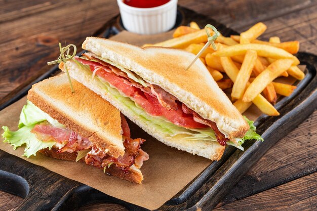 Club-Sandwich auf einem Holzbrett Neben Pommes Frites und einer Tasse Ketchup-Sauce Das Füllen des Sandwichs besteht aus Hähnchenbrust, Speck, Käse, Salat, Tomate, Nahaufnahme Makrofotografie