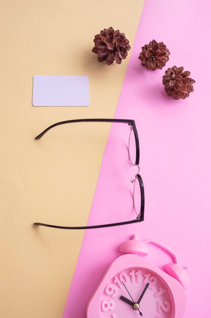 Clubmaster runde Brille in minimalem Sommerfoto auf pastellrosa und gelbem Hintergrund. Wecker , Kiefernblüten , Visitenkarten