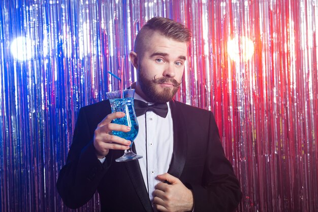 Clubparty- und Urlaubskonzept - Porträt eines eleganten, gutaussehenden Mannes in einem teuren Anzug hält blauen Cocktail auf glänzendem Hintergrund.