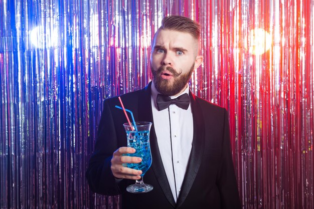 Clubparty- und Urlaubskonzept - Porträt eines eleganten, gutaussehenden Mannes in einem teuren Anzug hält blauen Cocktail auf glänzendem Hintergrund.