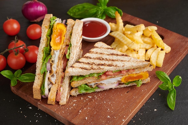 Foto clubsandwich com batatas fritas sanduíche com bacon ovo frito torrada grelhada e prensada com bacon ovo frito tomate e alface servido em tábua de madeira