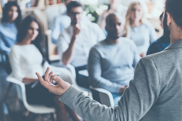 Coach de negocios. Vista trasera del hombre gesticulando con la mano mientras está de pie contra un grupo desenfocado de personas sentadas en las sillas frente a él