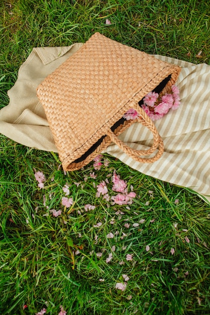 Cobertor de piquenique com chapéu de palha e bolsa na grama verde coberta com espaço de cópia de flores de sakura rosa