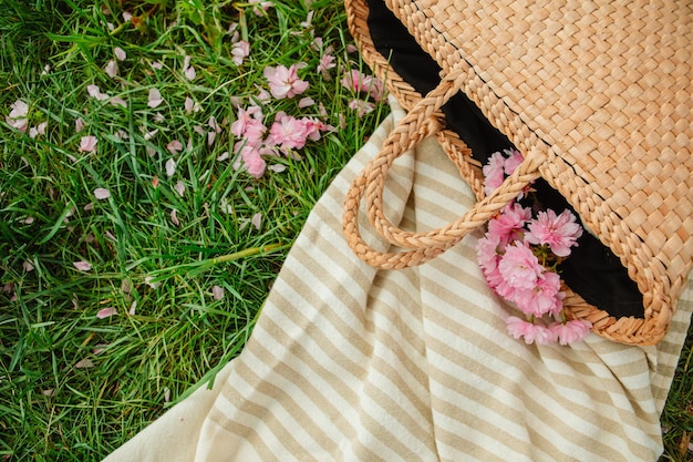 Cobertor de piquenique com chapéu de palha e saco em grama verde coberto com flores de sakura rosa