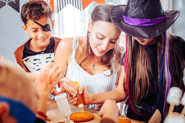 Cobertura em cookies. mãe carinhosa e amorosa ajudando seus filhos com rostos pintados e fantasias assustadoras colocando cobertura em biscoitos