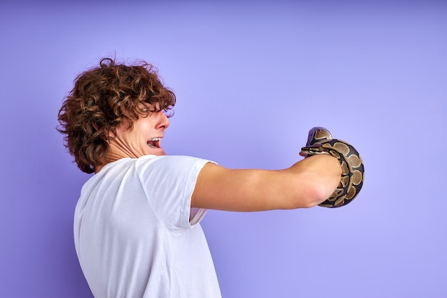 Foto cobra amarrada à mão, macho assustado está em choque, vista lateral em cara encaracolado olhando para o braço com cobra isolada no fundo roxo