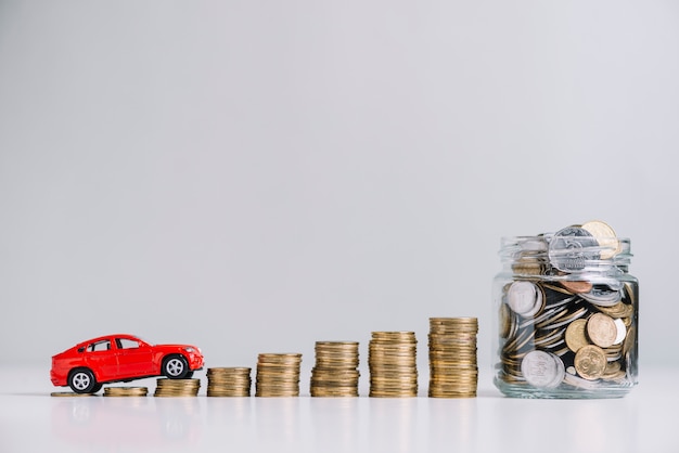 Coche conduciendo sobre el aumento de monedas apiladas cerca del tarro de cristal