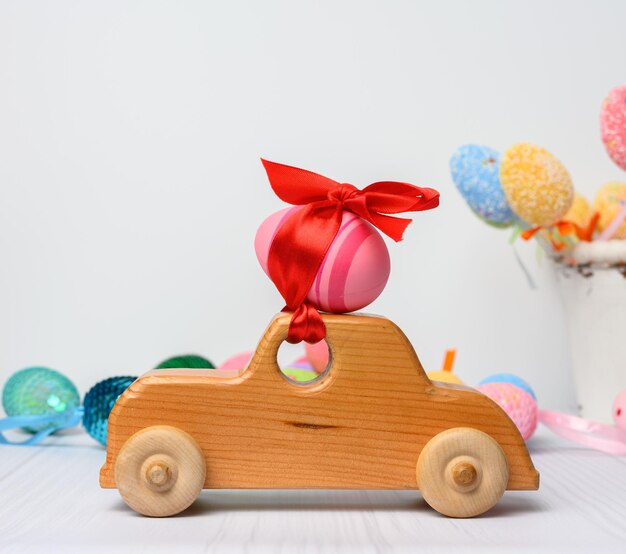 Foto coche de juguete de madera que lleva un huevo de pascua rosa fijado con un fondo blanco de cinta roja de cerca