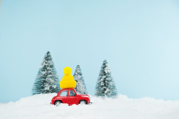 Coche rojo de Navidad con gorro amarillo tejido en un bosque de pinos nevados. Tarjeta de feliz año nuevo