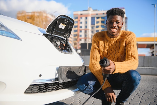 Coches eléctricos, concepto EV, combustible ecológico. Retrato de joven negro sonriente, recargando su coche eléctrico de lujo moderno