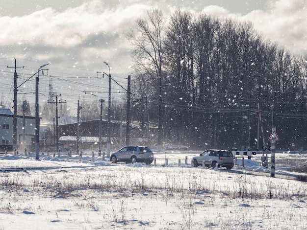 Los coches pasan por un cruce de ferrocarril fuera de la ciudad en invierno. Cruce ferroviario. Paisaje de invierno con árboles cubiertos de nieve y el sol.